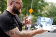A tattooed man with glasses is animatedly talking while holding a black-and-white photograph of a city street.