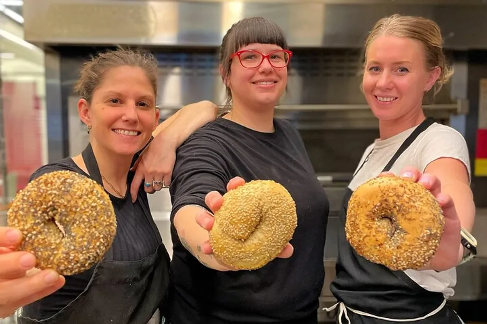 Three smiling individuals in a kitchen setting proudly hold up large seed-covered bagels towards the camera