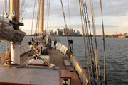 A view from the deck of a tall ship with its rigging visible, sailing towards a city skyline during sunset.