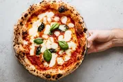 A hand is holding a freshly baked pizza topped with melted mozzarella, basil leaves, and a rich tomato sauce, with some charred spots on the crust indicating it may have been cooked in a wood-fired oven.