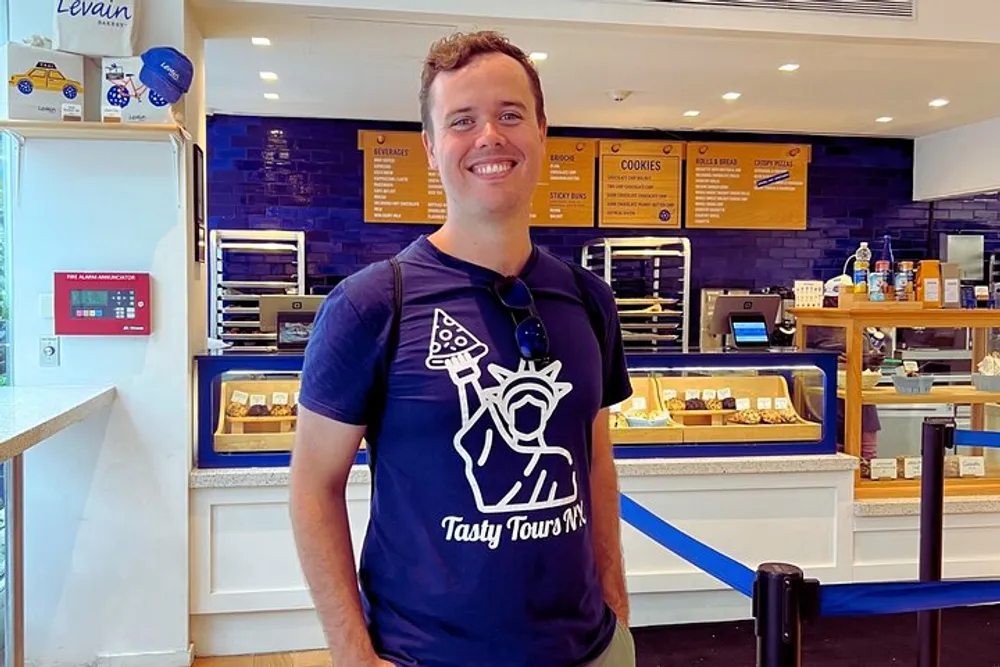 A smiling man stands inside a bakery with blue walls adorned with a Tasty Tours NY T-shirt in front of a counter displaying various baked goods