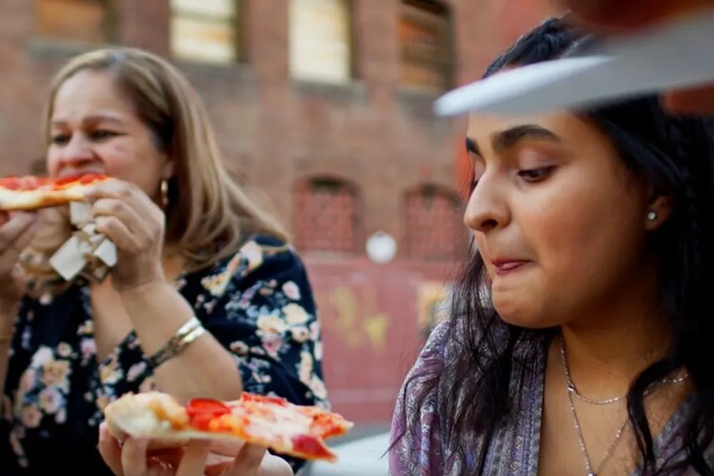 Two people are enjoying slices of pizza outdoors with one person in the foreground slightly out of focus as they gaze at a slice of pizza in their hand and the other person is in the background taking a bite