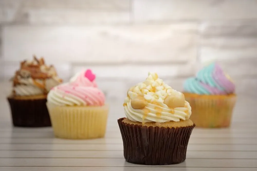 Four decorative cupcakes with various icing flavors are arranged in a row on a tabletop with a wooden background