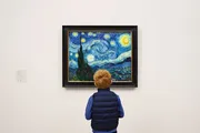A child is standing in front of and observing a framed painting of a starry night over a small town.