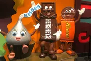 The image shows three anthropomorphic candy characters, representing Hershey's chocolate products, with a Kisses character on the left, a Hershey's bar in the center, and a Reese's package on the right, all with smiling faces and positioned as if greeting viewers.