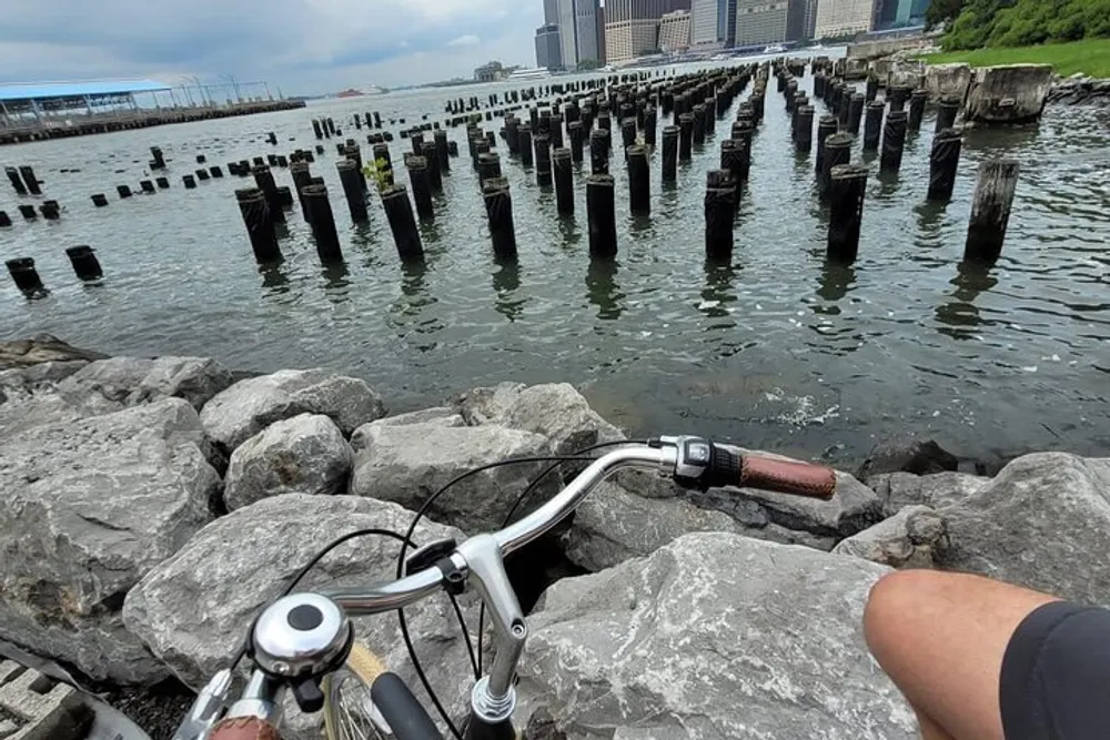 A cyclists perspective while resting on a rocky shore overlooking remnants of old piers jutting out of the water with a city skyline in the distance