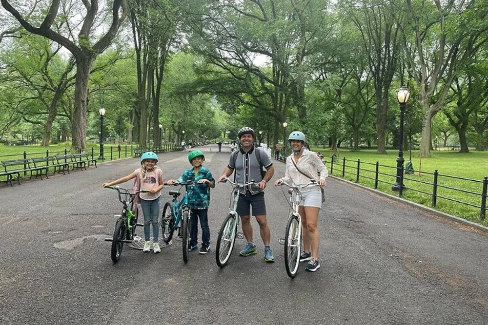 A family of four wearing helmets is standing with their bicycles on a tree-lined path in a park seemingly enjoying a day of bike riding together