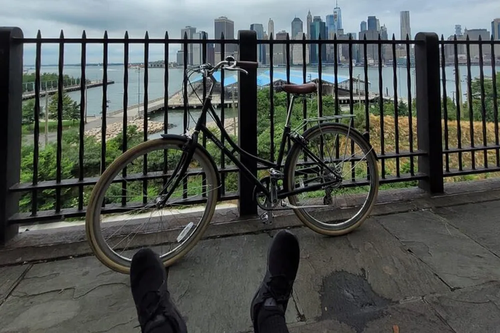 A person is taking a break and resting their feet with a view of a bike against a fence overlooking a river and a city skyline