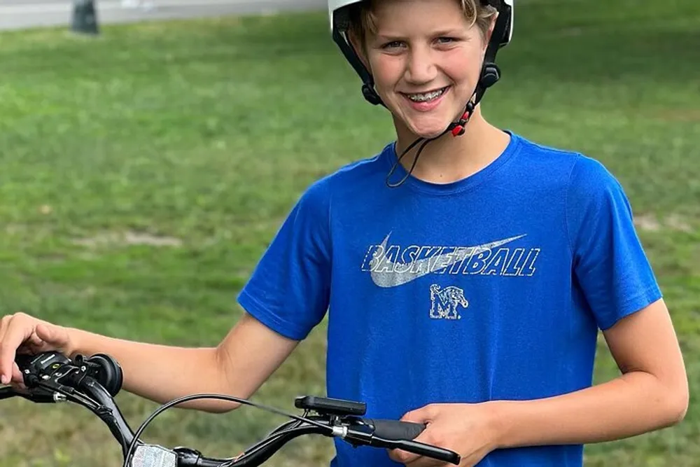 A smiling young person in a blue basketball t-shirt and a helmet is holding onto the handlebars of a bike outdoors