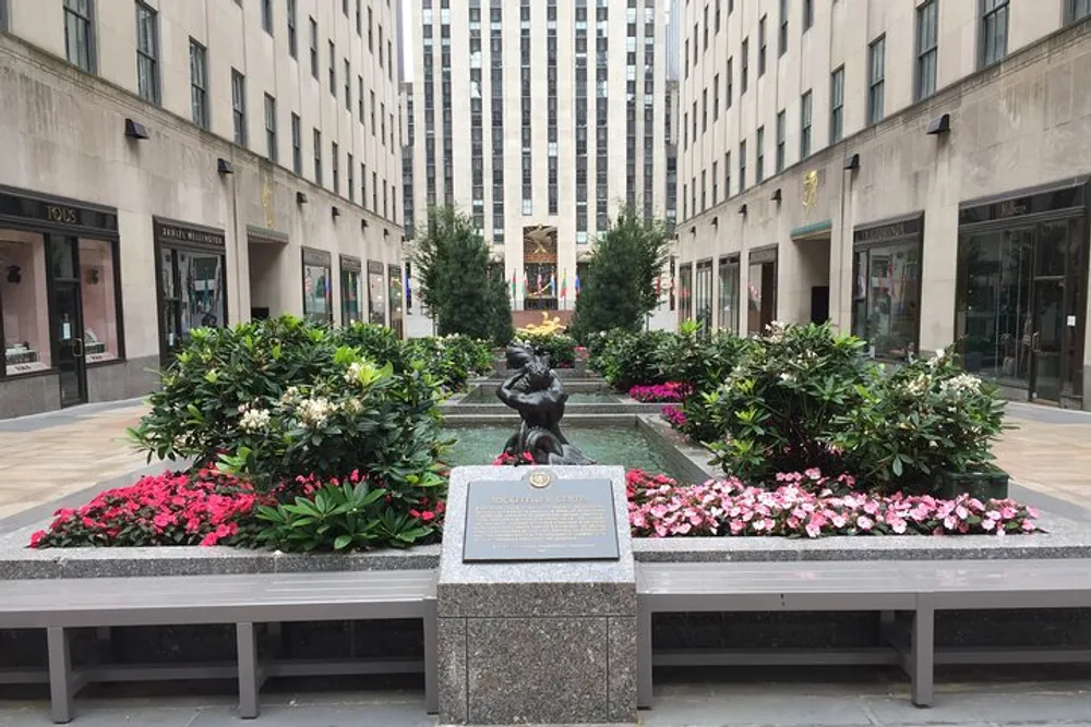 A peaceful urban plaza features a central statue surrounded by vibrant flowers flanked by benches and framed by tall buildings