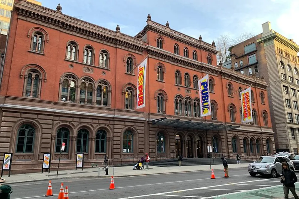 The image shows a large ornate red brick building with multiple arched windows and a series of colorful banners hanging from its facade situated on a city street with pedestrians and vehicles nearby