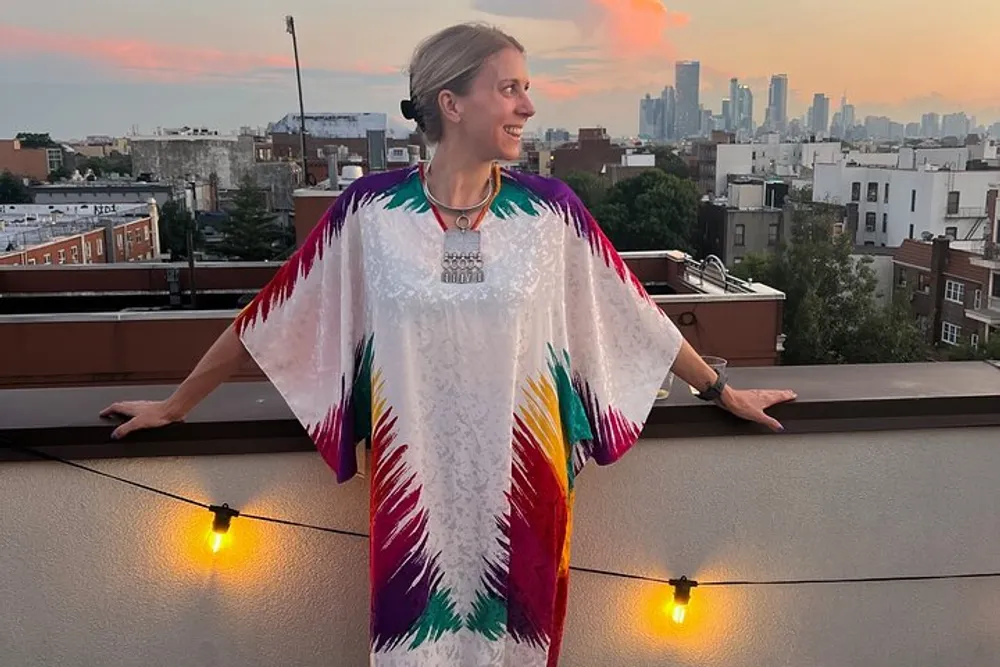 A smiling person is standing on a rooftop wearing a vibrant multicolored garment with a city skyline in the background during sunset