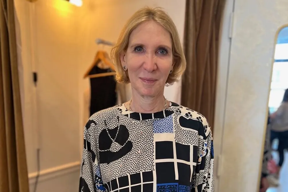 A woman with a pleasant expression is wearing a black and white patterned top and stands in a room with a mirror and a garment on a hanger in the background