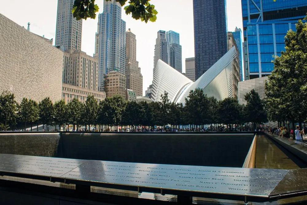 The photo shows a memorial with inscribed names in the foreground overlooking a reflecting pool with the distinct wing-like architecture of the Oculus in the background set against a cityscape of skyscrapers