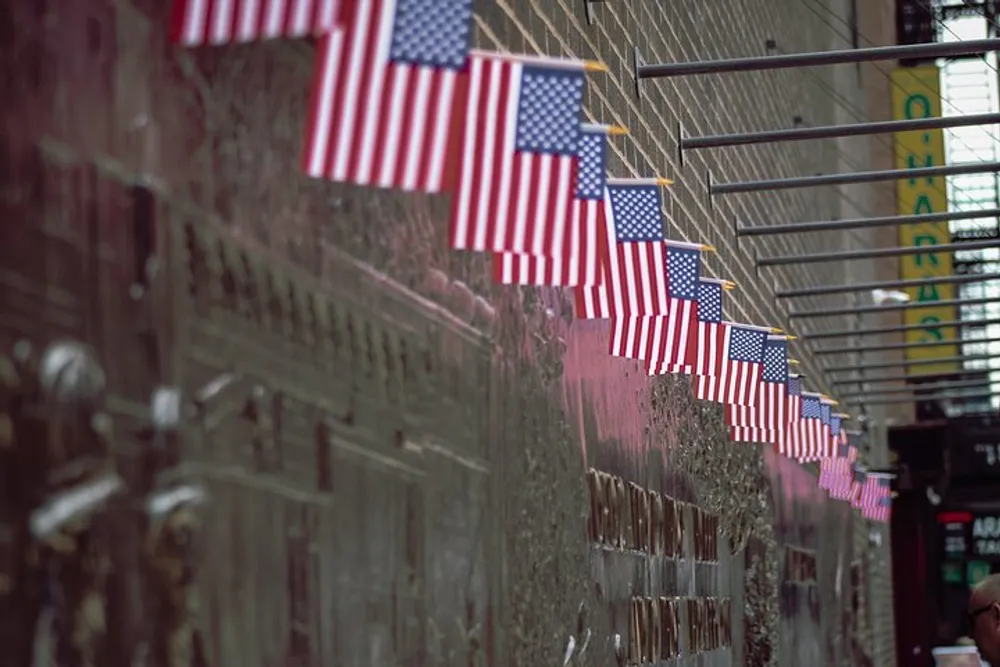 A series of small American flags are attached to a chain-link fence possibly as a form of patriotic display or commemoration