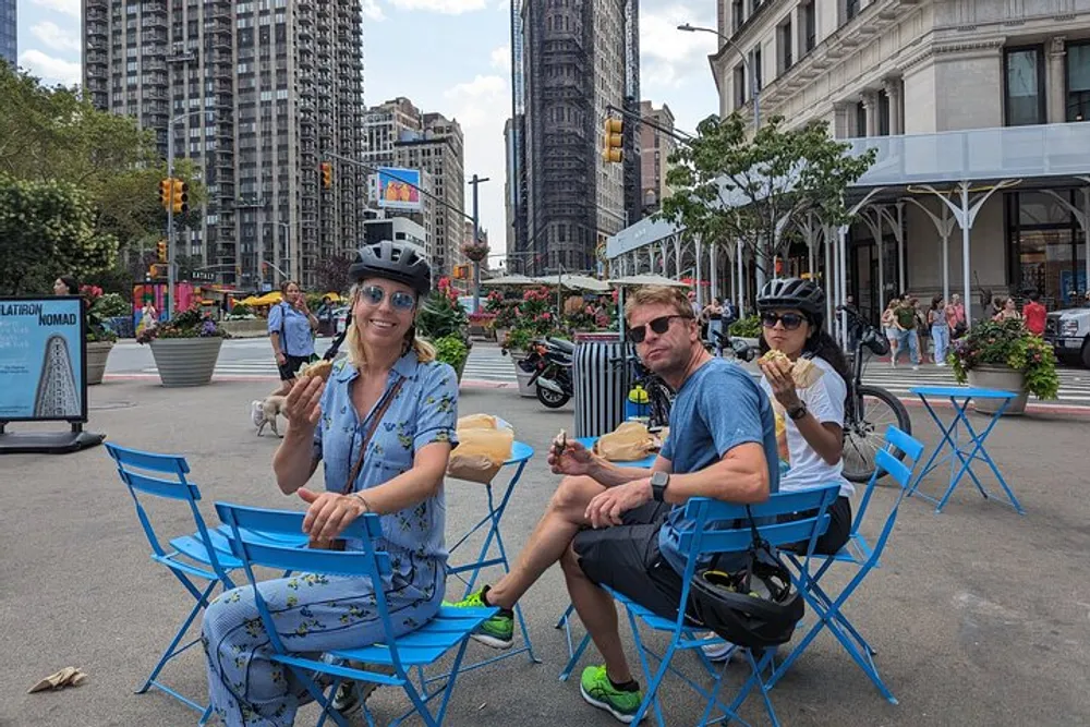 Three cyclists are taking a break with snacks at a blue outdoor public seating area in an urban environment with a tall narrow building in the background