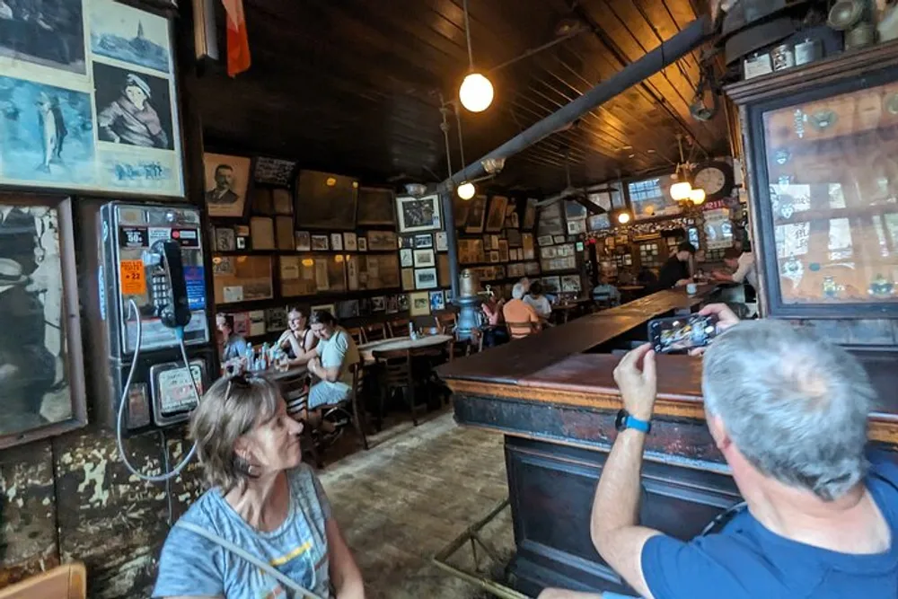 The image shows patrons inside a rustic bar with vintage decor and framed pictures on the walls as a man takes a photo of the setting