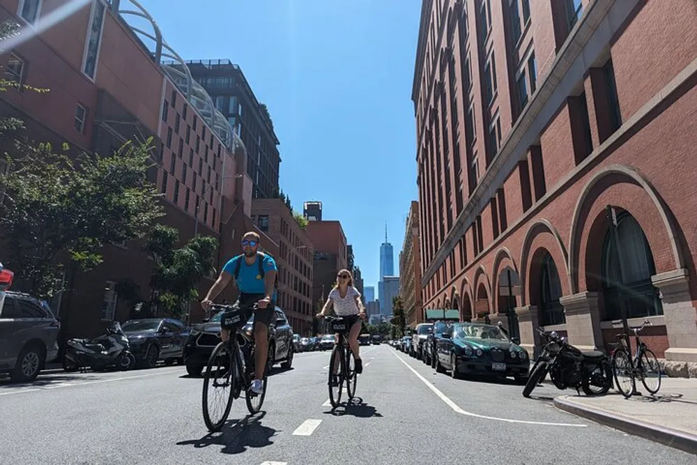 Two people are cycling down a sunlit urban street flanked by red brick buildings with a view of a tall skyscraper in the distance