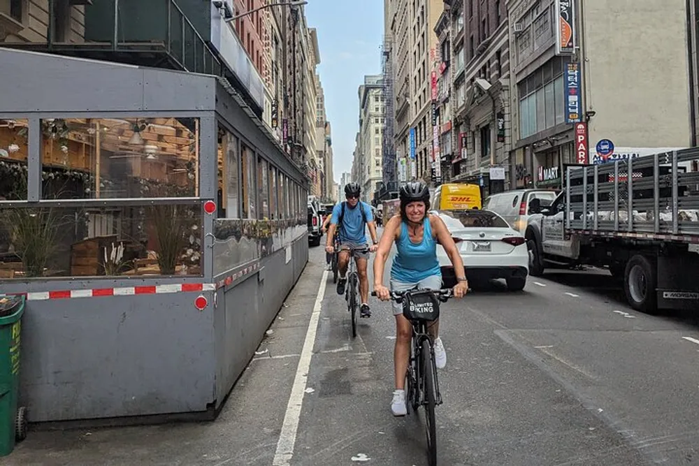 Two people are cycling down a city street with vehicles and buildings flanking them on both sides
