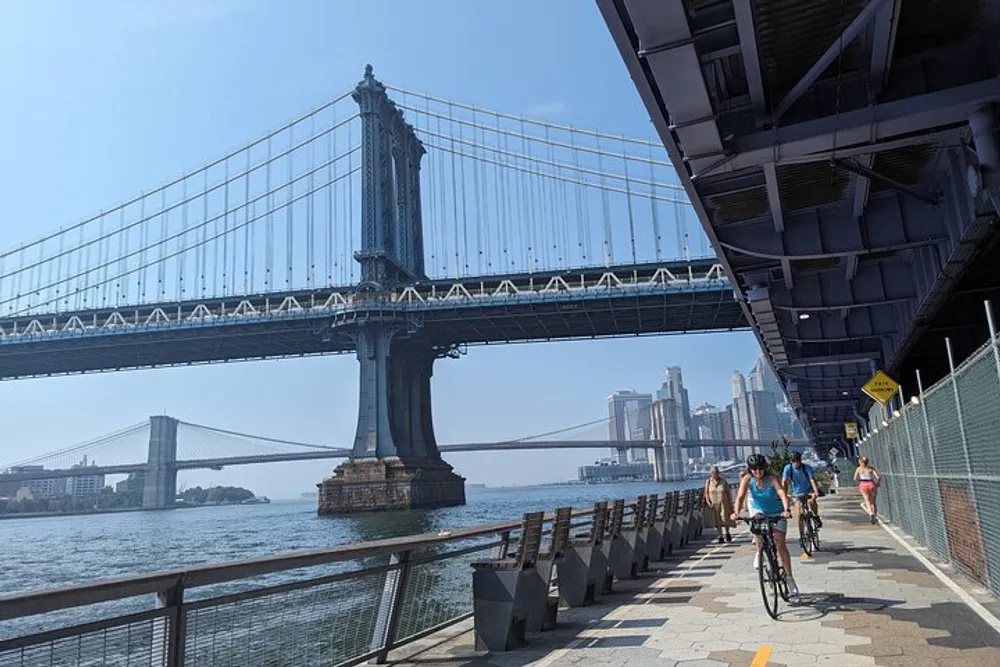 The image captures cyclists and pedestrians enjoying a sunny day on a path along the riverfront with a view of a grand suspension bridge in the background