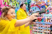 A smiling person in a yellow poncho and safety goggles is aiming a paint gun in front of a colorful graffiti-covered wall with another person in the background.