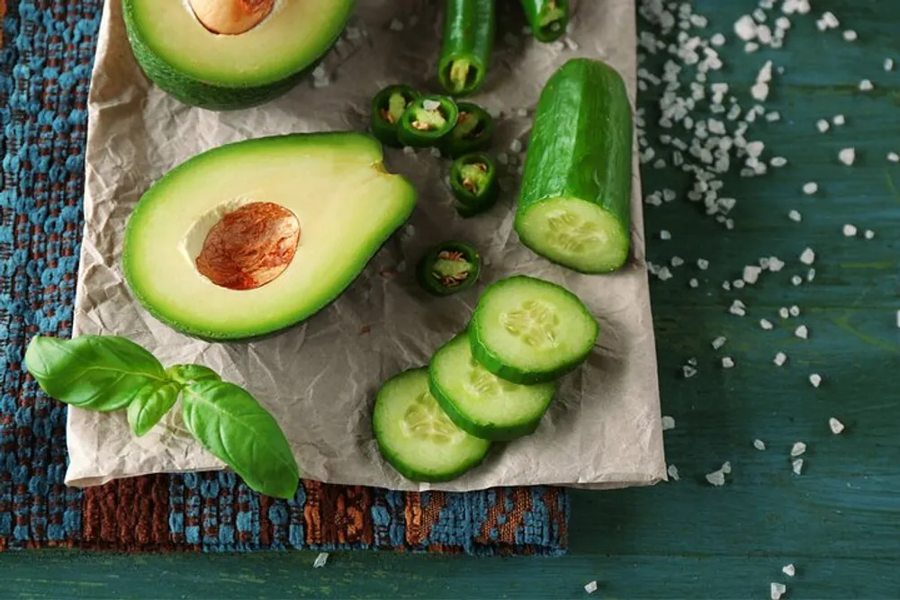 A halved avocado sliced cucumber jalapeo slices and basil leaves are scattered on parchment paper with salt crystals creating a fresh vegetable still life on a rustic wooden table