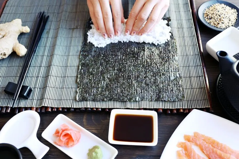 A person is preparing sushi with hands pressing down rice on a sheet of nori alongside various sushi ingredients and condiments