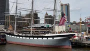 A large, traditional multi-masted sailing ship is moored in a modern urban harbor, flying the American flag among others.