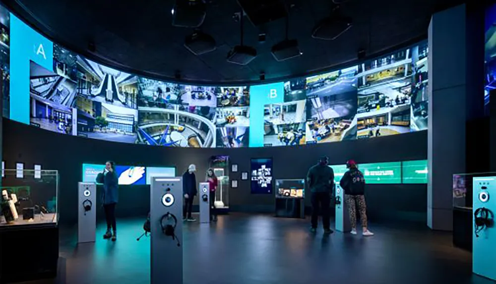 This image shows visitors engaging with interactive exhibits in a modern darkened room surrounded by large screens displaying surveillance camera footage