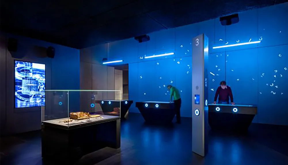 The image shows a modern dimly lit museum exhibition space with interactive displays and visitors engaging with the exhibits