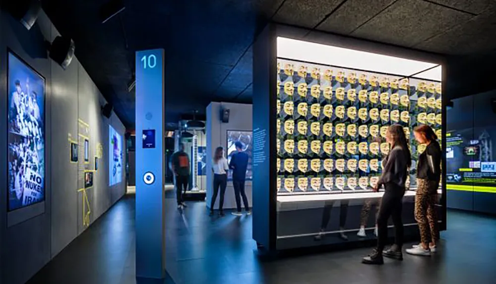 Visitors are viewing exhibits in a museum with a prominent display featuring rows of yellow masks against a lit background