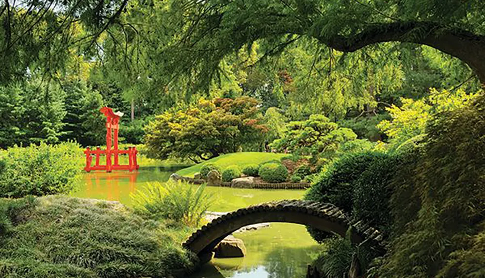 The image depicts a serene Japanese garden with a vibrant red torii gate on the water a stone arch bridge and lush greenery creating a tranquil setting