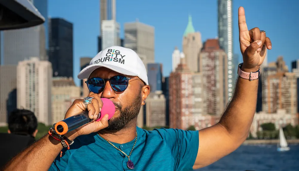 A person wearing sunglasses and a Big City Tourism cap is speaking into a microphone while gesturing with a raised index finger with a city skyline in the background