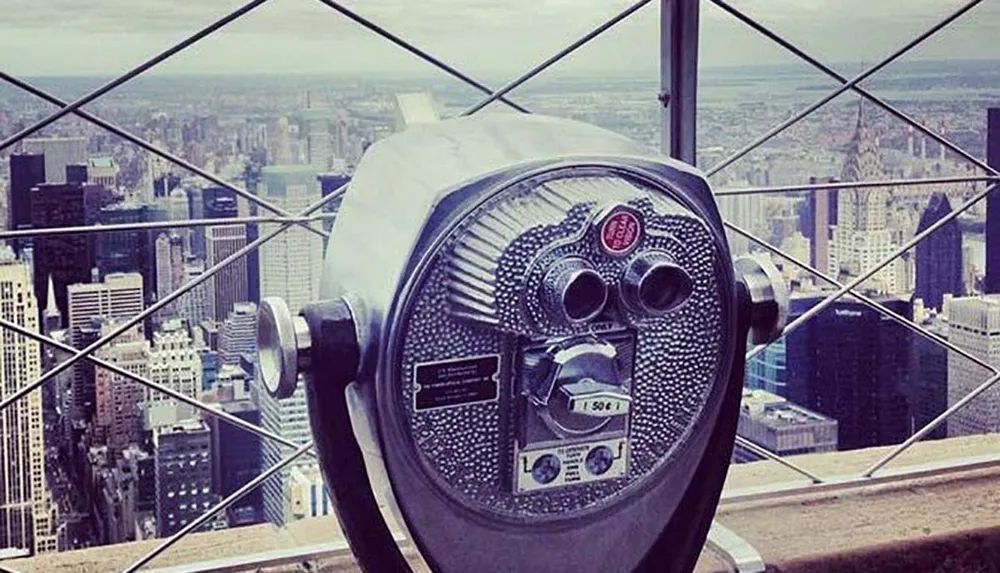 A coin-operated binocular viewer overlooking a cityscape from a high vantage point enclosed by safety railings