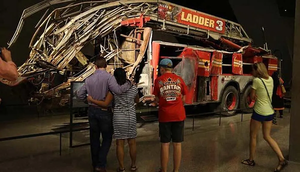 Visitors are observing a severely damaged firetruck on display that appears to be a remnant of a catastrophic event