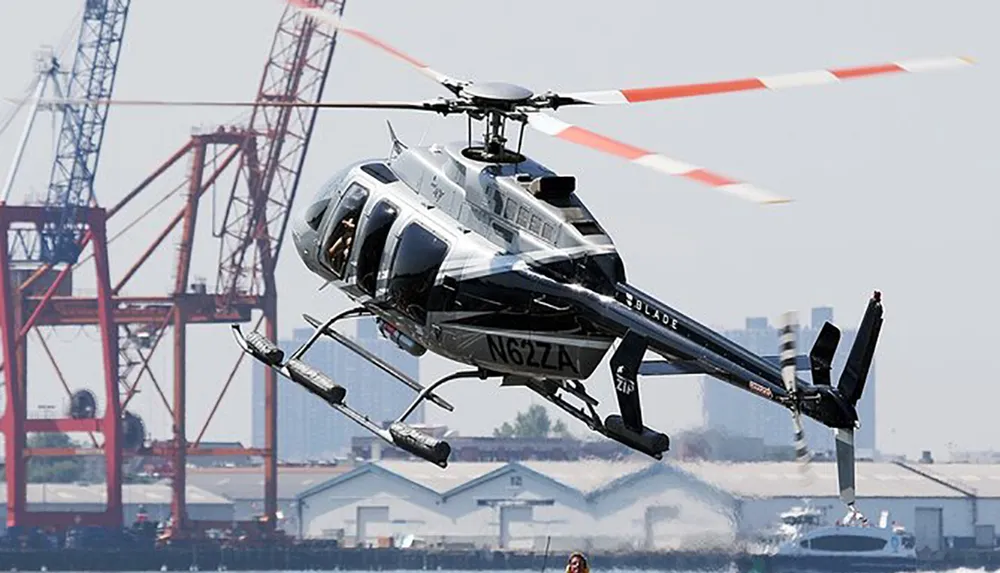 A helicopter equipped with water-dispensing equipment is in mid-flight against an industrial backdrop with cranes