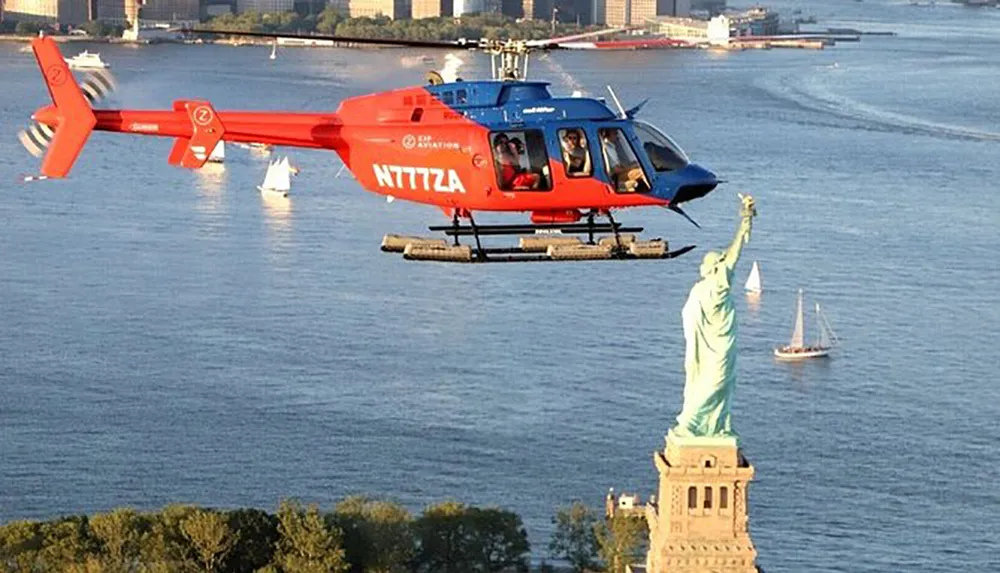 A red helicopter is flying near the Statue of Liberty with a backdrop of a calm sea dotted with boats
