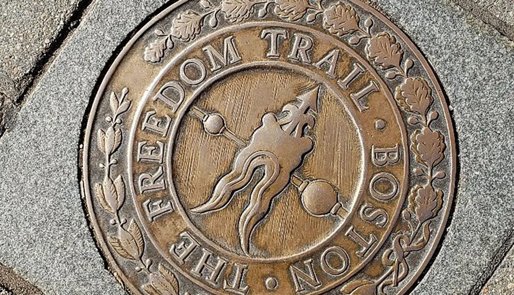 The image shows a bronze medallion embedded in the ground marking the iconic Freedom Trail in Boston adorned with an image of a revolutionary figure and surrounded by the inscription FREEDOM TRAIL BOSTON