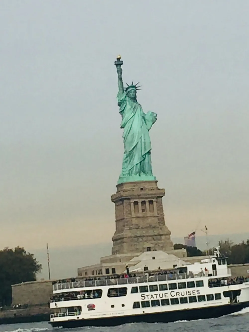 The image shows the iconic Statue of Liberty with a Statue Cruises ferry passing by in the foreground