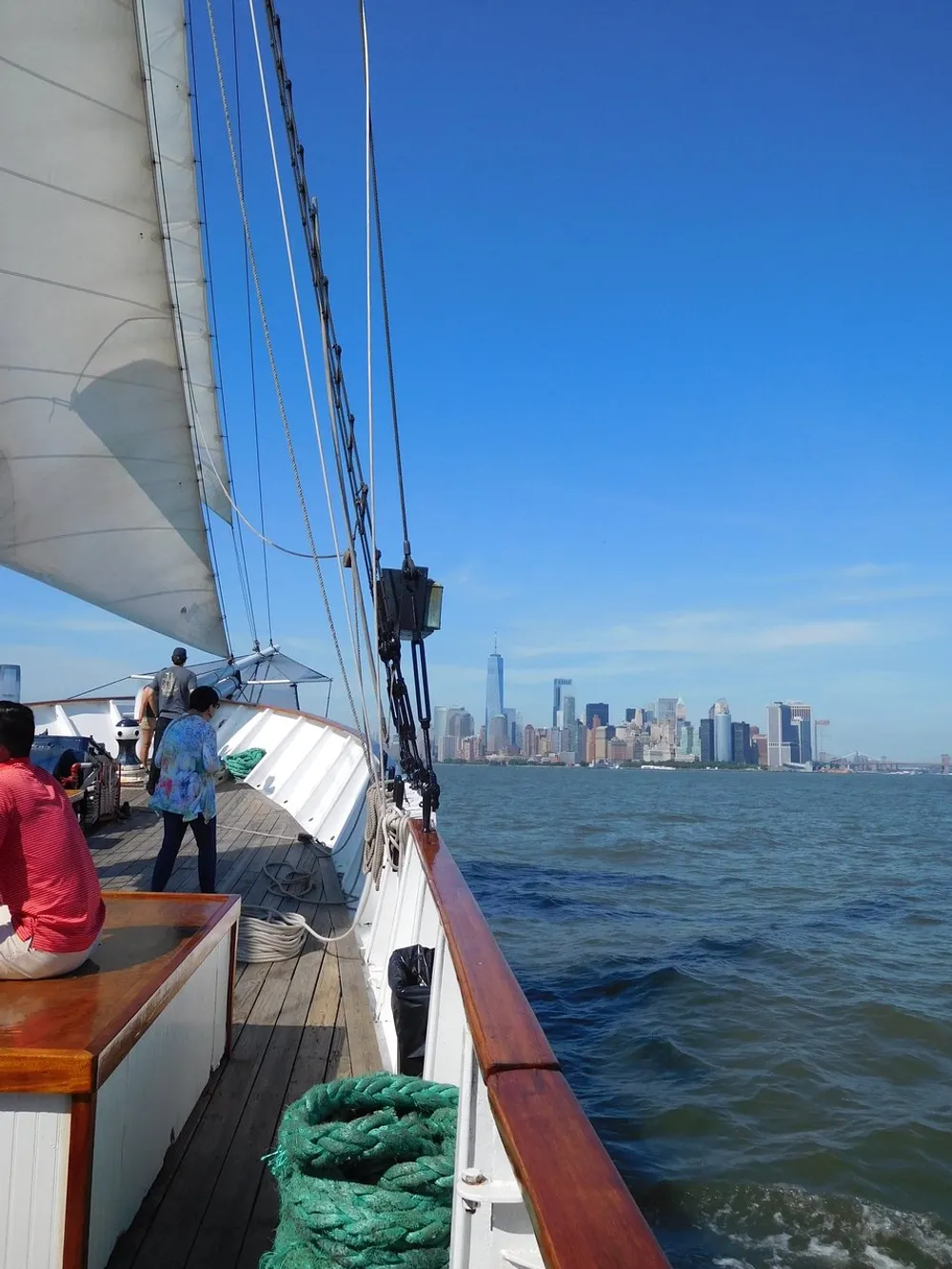 Passengers aboard a sailboat enjoy the views of a sprawling city skyline under a clear blue sky