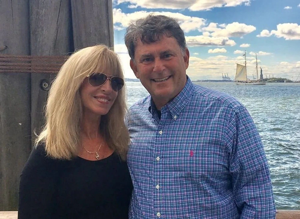 A smiling couple stands close together with a scenic background featuring the ocean and a sailboat under a partly cloudy sky