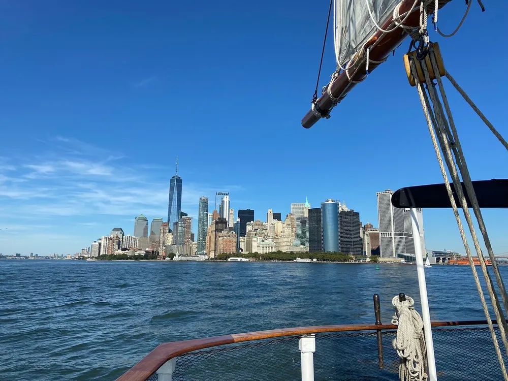 The image shows a view of the New York City skyline from a boat with the One World Trade Center standing prominently against a clear blue sky