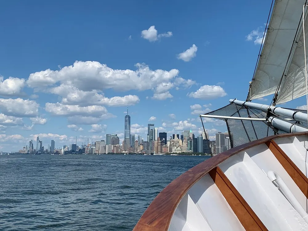 A sailboat is cruising near a bustling city skyline under a sky scattered with fluffy clouds