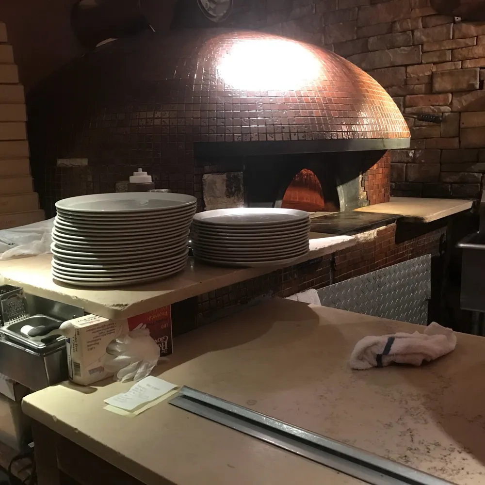 The image shows a wood-fired pizza oven with a glowing flame surrounded by piles of plates and kitchen utensils suggestive of a restaurant kitchen preparing for service