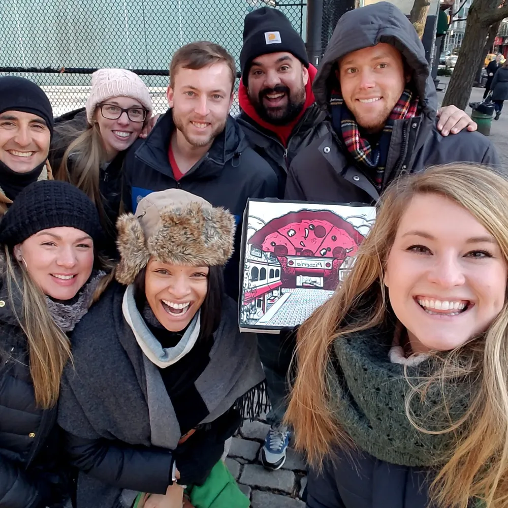A happy group of seven people in winter attire pose for a selfie with one person holding a colorful artwork