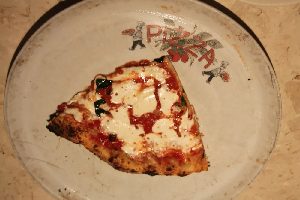 A single slice of pizza with tomato sauce cheese and what looks like basil rests on a decorative plate with the word PIZZA and an illustration of a chef