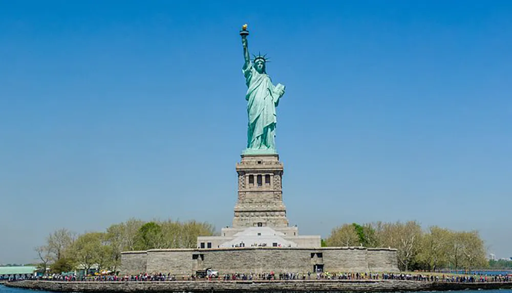 The image shows the Statue of Liberty standing tall against a clear blue sky with numerous visitors visible at its base