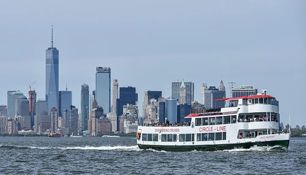 A Circle Line sightseeing cruise boat is traveling along the water with the New York City skyline including One World Trade Center in the background