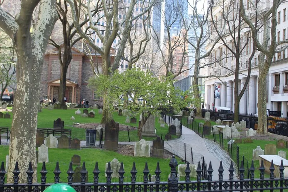 The image shows a historical cemetery with weathered headstones nestled among trees and pathways surrounded by a wrought iron fence with city buildings as a backdrop