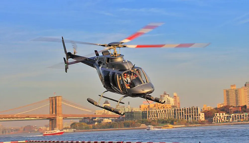 A helicopter is flying near a bridge over a river in an urban setting with two people visible inside